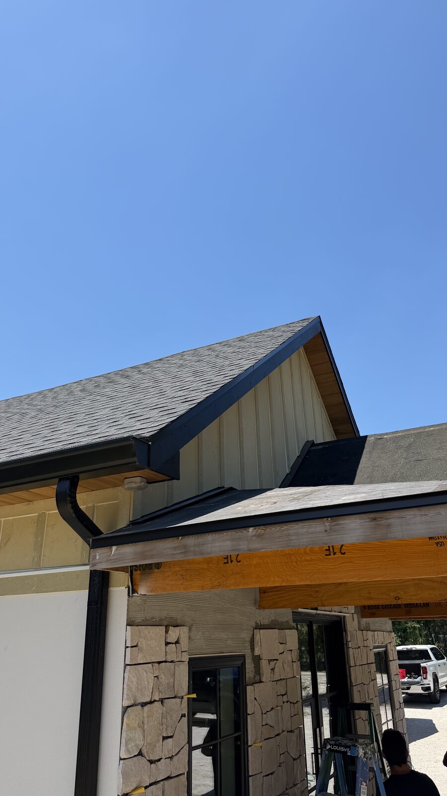 Black fascia and downspout wrapping a gable with vertical metal siding above a stone covered entry